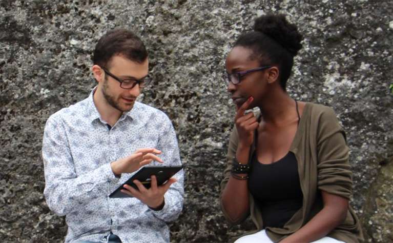 A man and woman have a discussion while the man scrolls on a mobile tablet.