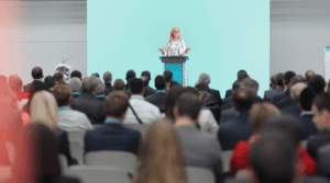 A blonde female doctor in a white coat speaks in front of an audience at a conference.