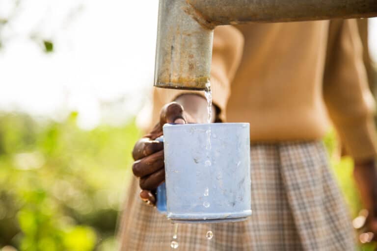 An African woman filling water in a blue metal mug from a rusty sink in a village.