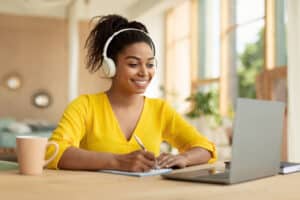 A woman in a yellow top and headphones is working at a laptop.